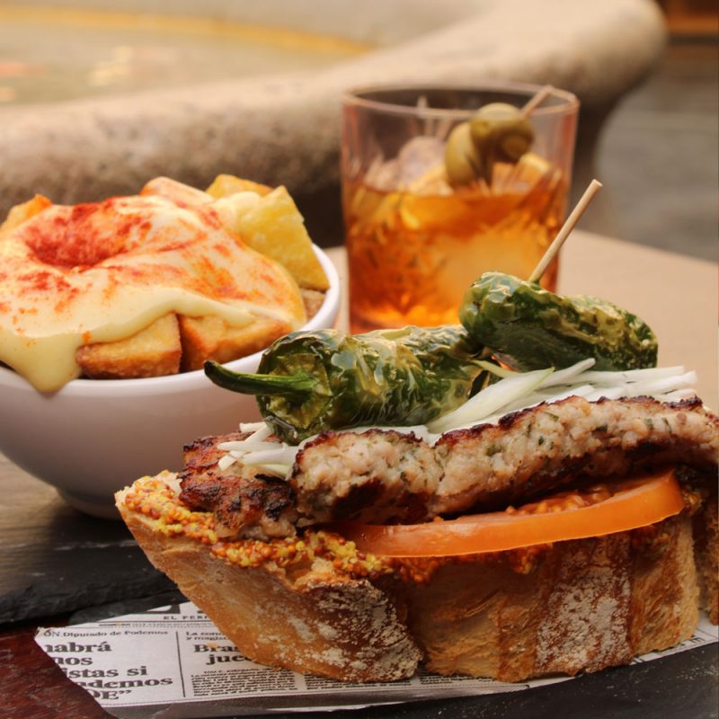 Nuestro montadito del longaniza casera a las finas hierbas, tomate, cebolla tierna, mostaza y pimiento de padrón.